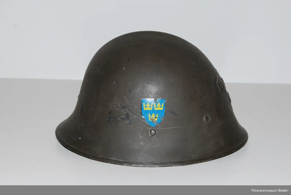 Mörkt grågrön stålhjälm av typen med smalare och högre brätte, den till höger på bild 6 som jämför båda typerna av hjälm m/1921. Med insats bestående av tre läderkuddar som hålls ihop med skorem. Hakband av läder. Framtill tre kronor i relief, tre-kronor dekaler på sidorna. Upptill en förstärkningspressning i form av en kam.
