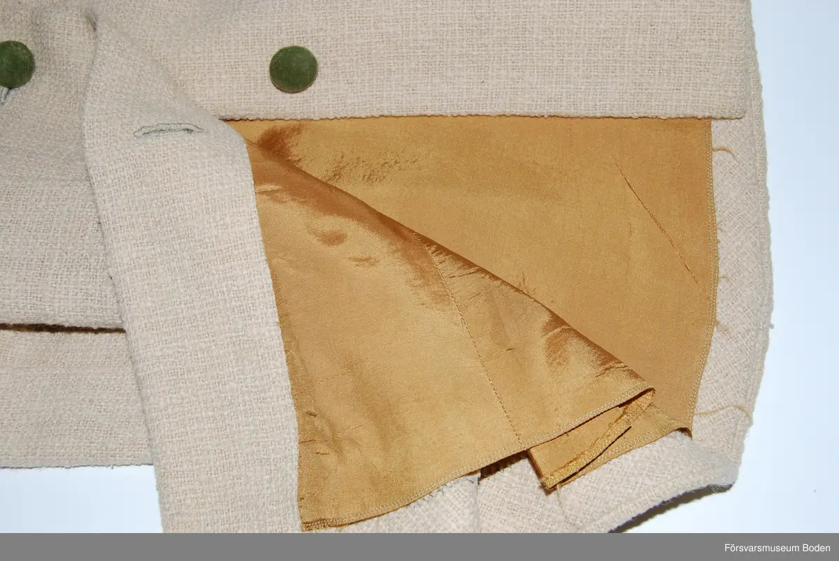 Beige textil med knappar och kragfoder av grön plysch. Kappans foder av senapsgult sidentyg. Ej fabrikstillverknad, saknar märkning.