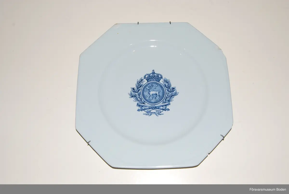 Flat 8-kantig tallrik av blåtonat porslin (flintgods) av Rörstrands tillverkning. I centrum en blå vapenbild med Västerbottens landskapsbild i rund, krönt sköld omgiven av lagerkrans. Nertill under skölden korslagda eldrör och knippe med blixtar.
