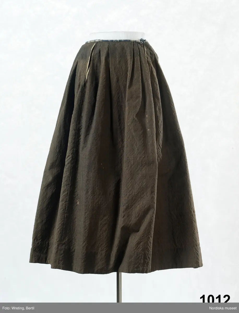 Kjol av gråsvart strävt och glansigt kamgarnstyg s.k. kamlott, veckad med motveck mitt bak, sprund vid vardera sidan om framvåden som har 3 veck åt vardera sidan, slät fram. Linning och 8 cm kantskoning av blå- och vitrutig linnelärft.

Använd som brudkjol midsommardagen 1795 av Annika Andersdotter. Hela bruddräkten inv.nr 1012 -1018. Även brudgummens dräkt bevarad inv.nr 1019- 1021. Se bilder i Anna-Maja Nyle´n: Folkdräkter, 1971.
Berit Eldvik okt 2005