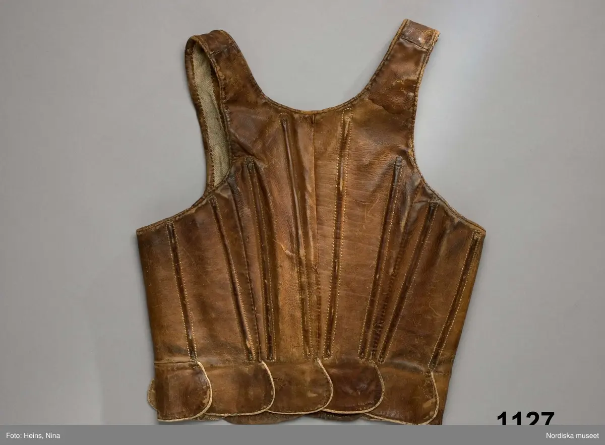 Katalogkort:  Lifstycke af brunt kalvskinn. Snörlif med insydda styfva spröt i rygg, sidor och framtill, sex par snörhål och en snörrem af skinn i det nedersta hålet på höger sida samt tio tegellagda flikar delvis i ett stycke med lifvet, delvis tillskarfvade. Foder af grov hvit linnehväf.
Tillhör den äldre kvinnodräkten, kalladt "munkalifstycke". Dylika lif, men af rödt kläde och med grönkantade flikar, förekommo i bruk långt in på 1840-talet.
/P.G.Vistrand/

Skinnet brunt, kraftigt. Med sammanlagt 12 insydda spröt  jämnt fördelade runt livet., snörhålen kantsydda med ljust lingarn. Foder av oblekt linne i spetskypert.
Anm. Helt men väl använt, nött och fläckigt.
Sådana skinnlivstycken användes framförallt till vardags, de var mycket slitstarka.
/Berit Eldvik 2008-02 15Kalvskinn.