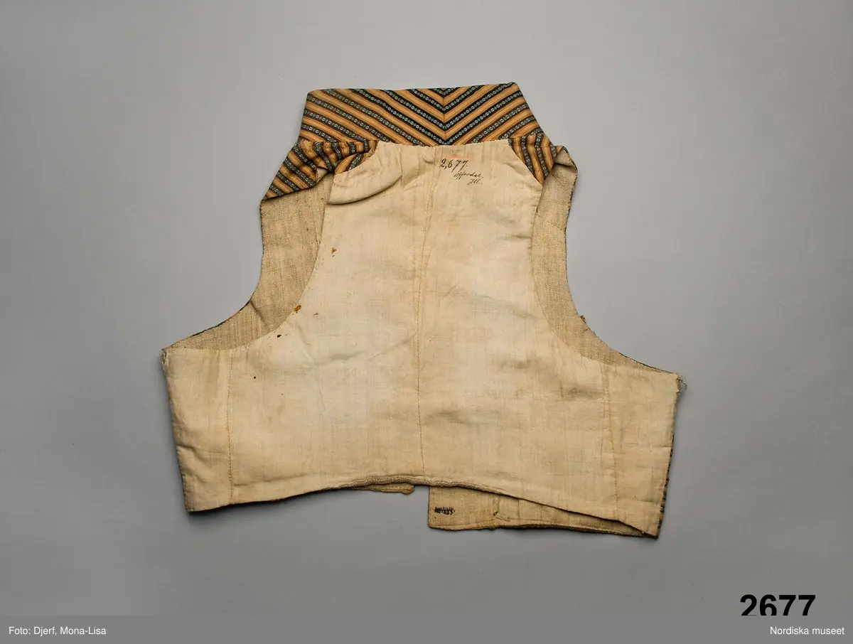 Väst i kort modell av randig bomullsrips i brunt och beige med invävd mönsterrand i den mörkbruna randen i ljusblått med vita "hoppor". Västen är tvärrandig med 2 framstycken och hög styvad  krage av samma tyg, ryggstycke av fin oblekt linnelärft, foder av grov och gles linnelärft i ryggen och finare i framstyckena, 2 stolpfickor, dubbelknäppning med knapphål på båda framstyckena och 6 par mässingsknappar med Ångermanlands landskapsvapen med 3 liggande laxar.
Tygsorten finns under benämningen: Västtyg i flera av 1820-30 talets tygprovsböcker och har enligt uppgift använts som brudgumsväst 1824
/Berit Eldvik  2005-11