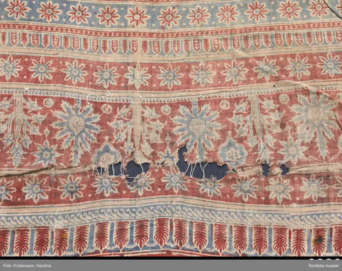 Stort tygstycke sammansytt av 3 våder bomullslärft  á 54 cm bredd. Hopsydda med vitt handspunnet lingarn. Tryckt mönster i rött och blått på vitt underlag.. I mitten stort cirkelmotiv fylld med mönster och i mitten en stjärna, åt fyra håll en triangel ut från cirkeln och mot 4 större snäckliknande ornament. I varje hörn motiv med 4 fiskar med stjärtarna in mot en cirkel, Kantbårder bestånde av olikmönstrade ränder i olika bredder.  Trycket är gjort med tryckstockar och smalare tryckstämplar i kantbårderna där tydliga blockskarvar syns.  Tekniken är den gamla indiska med betmålning av de partier som skall bli röda och därefter doppning i krappbad, tvättning av tyget före blåfärgning som görs så att vita och röda parteier täcks med en pasta före färgning i indigokyp. På baksidan är tyget mycket blåare än på framsidan. Spegelns botten har färgats först röd och därefter blå  så att den fått en mörklila , lätt flammig nyans. Tyget är skört och trasigt på flera ställen, längs hela högersidan är det söndigt vilket kan vara musätet. En del mindre hål har lappats med  gråblått siden på baksidan eller på ett ställe med en bit av själva tygtrycket. Även blekt.
Insamlat av Nils Månsson-Mandelgren vid hans resor i Västergötland, inga uppgifter finns.
 /Berit Eldvik 2013-06