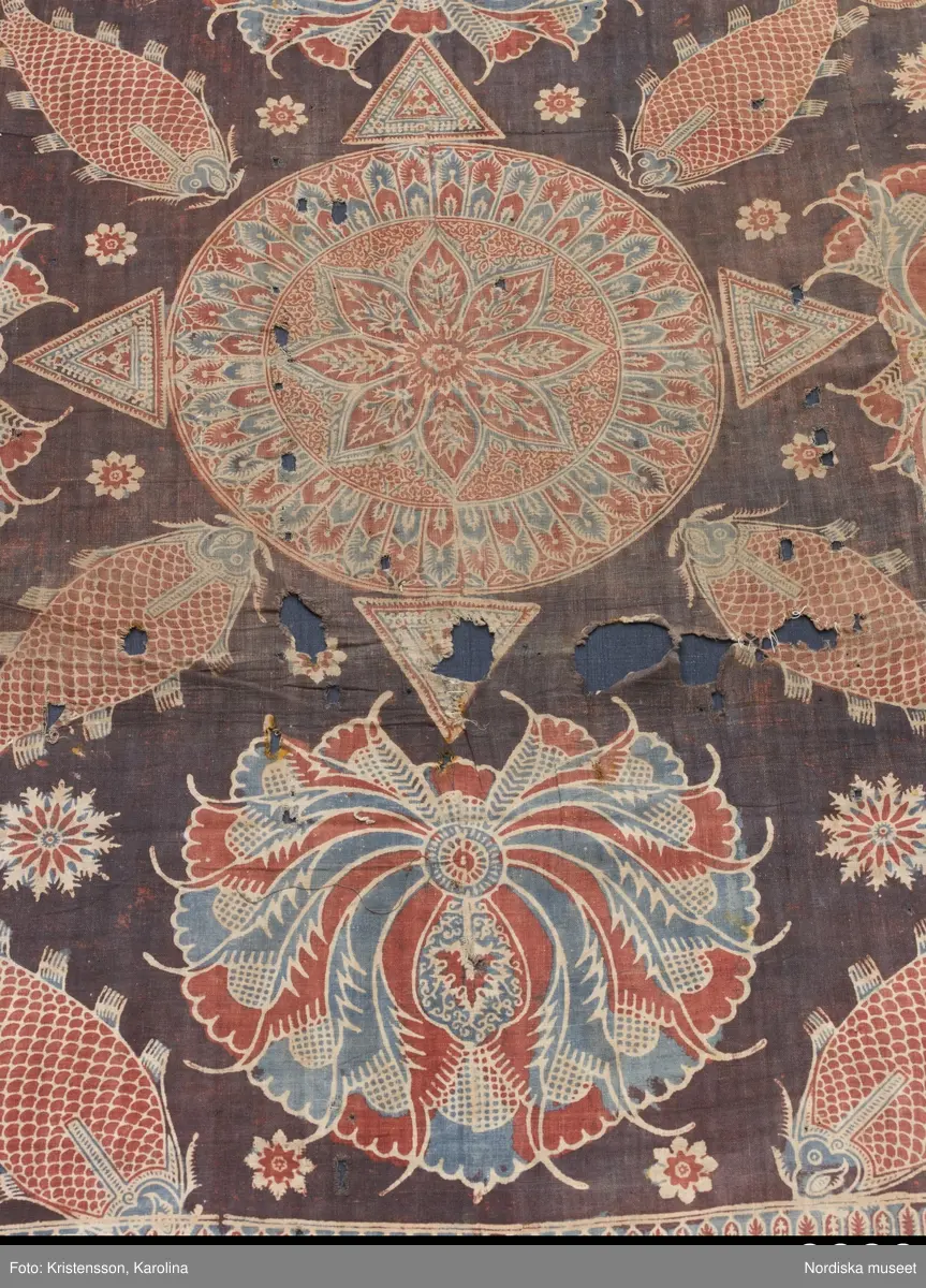 Stort tygstycke sammansytt av 3 våder bomullslärft  á 54 cm bredd. Hopsydda med vitt handspunnet lingarn. Tryckt mönster i rött och blått på vitt underlag.. I mitten stort cirkelmotiv fylld med mönster och i mitten en stjärna, åt fyra håll en triangel ut från cirkeln och mot 4 större snäckliknande ornament. I varje hörn motiv med 4 fiskar med stjärtarna in mot en cirkel, Kantbårder bestånde av olikmönstrade ränder i olika bredder.  Trycket är gjort med tryckstockar och smalare tryckstämplar i kantbårderna där tydliga blockskarvar syns.  Tekniken är den gamla indiska med betmålning av de partier som skall bli röda och därefter doppning i krappbad, tvättning av tyget före blåfärgning som görs så att vita och röda parteier täcks med en pasta före färgning i indigokyp. På baksidan är tyget mycket blåare än på framsidan. Spegelns botten har färgats först röd och därefter blå  så att den fått en mörklila , lätt flammig nyans. Tyget är skört och trasigt på flera ställen, längs hela högersidan är det söndigt vilket kan vara musätet. En del mindre hål har lappats med  gråblått siden på baksidan eller på ett ställe med en bit av själva tygtrycket. Även blekt.
Insamlat av Nils Månsson-Mandelgren vid hans resor i Västergötland, inga uppgifter finns.
 /Berit Eldvik 2013-06