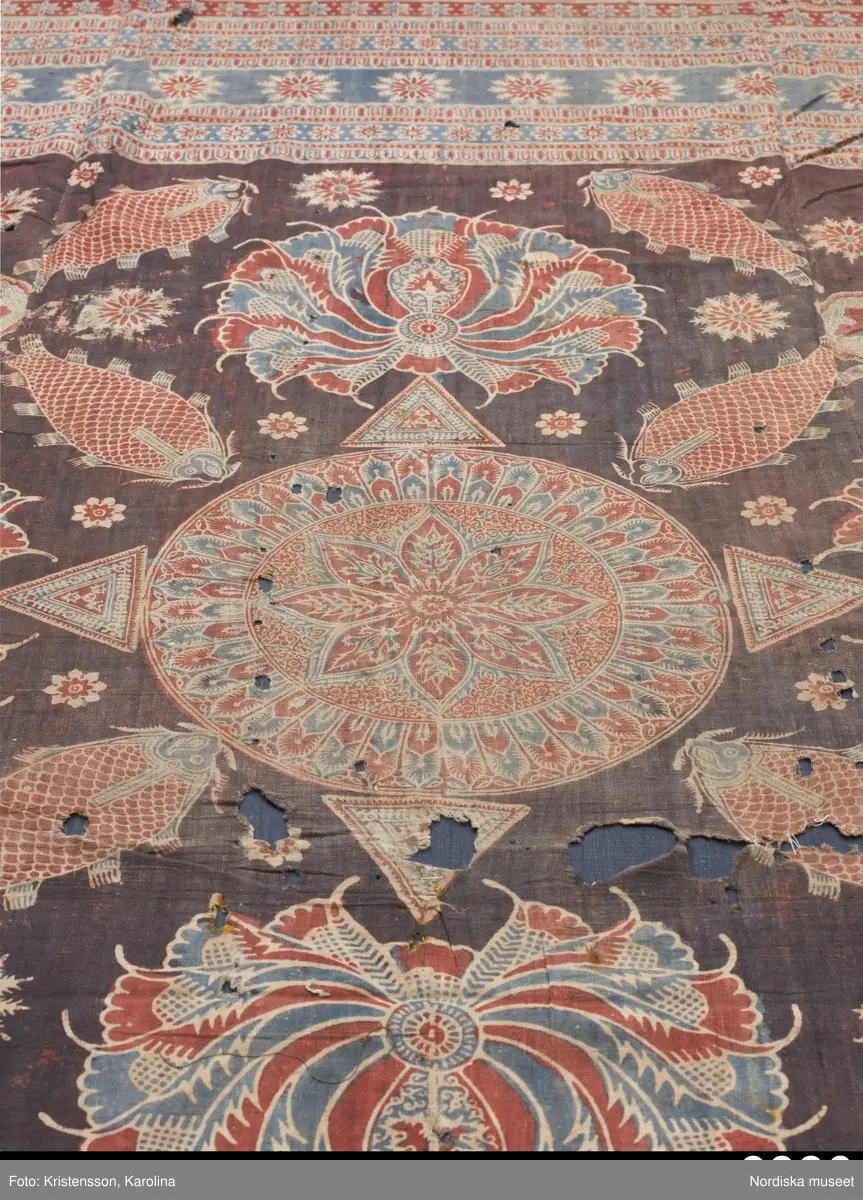 Stort tygstycke sammansytt av 3 våder bomullslärft  á 54 cm bredd. Hopsydda med vitt handspunnet lingarn. Tryckt mönster i rött och blått på vitt underlag.. I mitten stort cirkelmotiv fylld med mönster och i mitten en stjärna, åt fyra håll en triangel ut från cirkeln och mot 4 större snäckliknande ornament. I varje hörn motiv med 4 fiskar med stjärtarna in mot en cirkel, Kantbårder bestånde av olikmönstrade ränder i olika bredder.  Trycket är gjort med tryckstockar och smalare tryckstämplar i kantbårderna där tydliga blockskarvar syns.  Tekniken är den gamla indiska med betmålning av de partier som skall bli röda och därefter doppning i krappbad, tvättning av tyget före blåfärgning som görs så att vita och röda parteier täcks med en pasta före färgning i indigokyp. På baksidan är tyget mycket blåare än på framsidan. Spegelns botten har färgats först röd och därefter blå  så att den fått en mörklila , lätt flammig nyans. Tyget är skört och trasigt på flera ställen, längs hela högersidan är det söndigt vilket kan vara musätet. En del mindre hål har lappats med  gråblått siden på baksidan eller på ett ställe med en bit av själva tygtrycket. Även blekt.
Insamlat av Nils Månsson-Mandelgren vid hans resor i Västergötland, inga uppgifter finns.
 /Berit Eldvik 2013-06