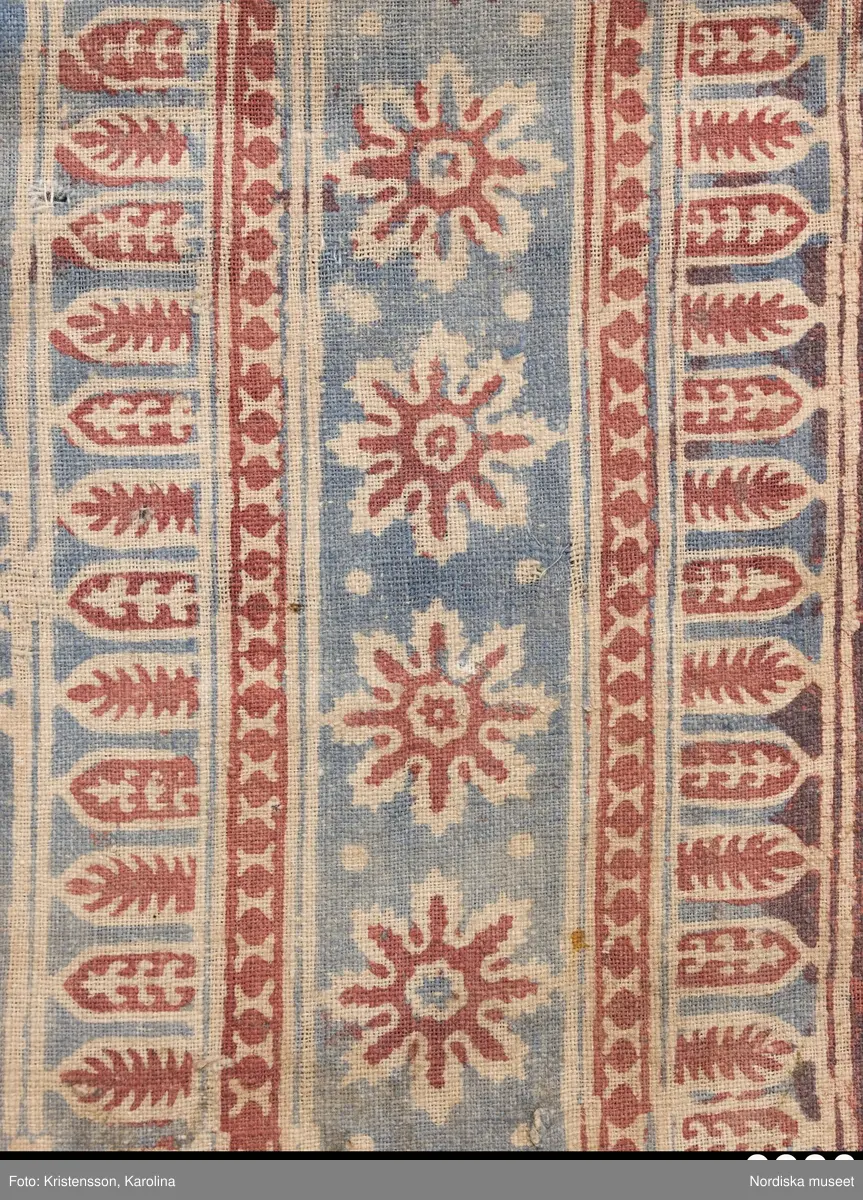 Stort tygstycke sammansytt av 3 våder bomullslärft  á 54 cm bredd. Hopsydda med vitt handspunnet lingarn. Tryckt mönster i rött och blått på vitt underlag.. I mitten stort cirkelmotiv fylld med mönster och i mitten en stjärna, åt fyra håll en triangel ut från cirkeln och mot 4 större snäckliknande ornament. I varje hörn motiv med 4 fiskar med stjärtarna in mot en cirkel, Kantbårder bestånde av olikmönstrade ränder i olika bredder.  Trycket är gjort med tryckstockar och smalare tryckstämplar i kantbårderna där tydliga blockskarvar syns.  Tekniken är den gamla indiska med betmålning av de partier som skall bli röda och därefter doppning i krappbad, tvättning av tyget före blåfärgning som görs så att vita och röda parteier täcks med en pasta före färgning i indigokyp. På baksidan är tyget mycket blåare än på framsidan. Spegelns botten har färgats först röd och därefter blå  så att den fått en mörklila , lätt flammig nyans. Tyget är skört och trasigt på flera ställen, längs hela högersidan är det söndigt vilket kan vara musätet. En del mindre hål har lappats med  gråblått siden på baksidan eller på ett ställe med en bit av själva tygtrycket. Även blekt.
Insamlat av Nils Månsson-Mandelgren vid hans resor i Västergötland, inga uppgifter finns.
 /Berit Eldvik 2013-06