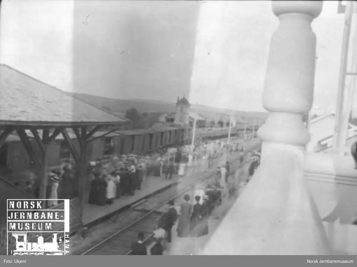 Oversiktsbilde med reisende på plattformene, et godstog og lokomotivstallen i bakgrunnen
