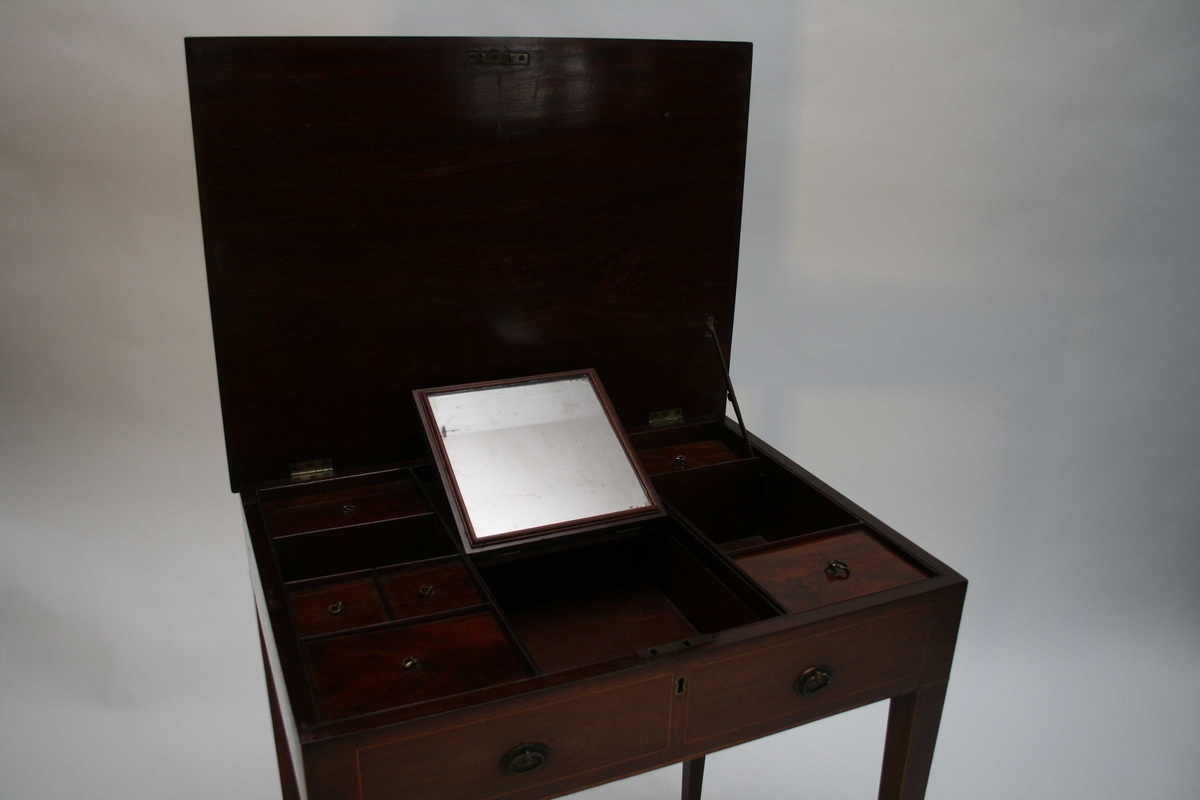 Polert mahognibord med intarsia. Rektangulær plate, avsmalende bein og to falske skuffer i sargen. Bordplata kan slås opp, og under er det et lite skyvespeil og 9 små rom, 6 rom har lokk.
