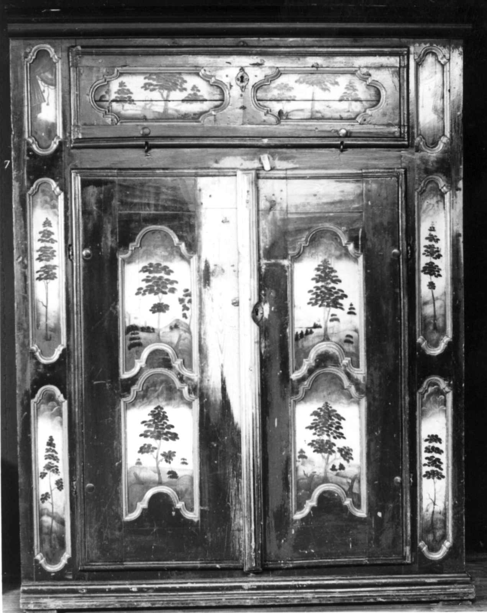 Flatskap med doble dører og klaff i overkant, hengslet på undersiden. Dekormalt med landskapsmotiver i feltene.