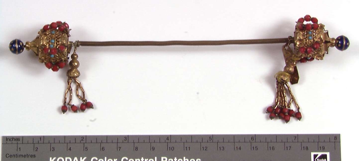 Hattenål i metall, delvis forgylt. Endestykkene er dekorert med røde og blå glassperler og dusker.