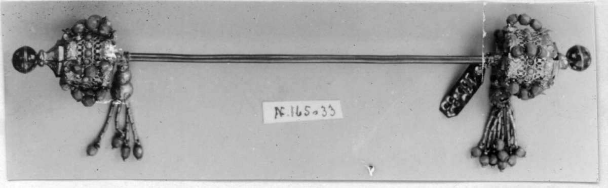 Hattenål i metall, delvis forgylt. Endestykkene er dekorert med røde og blå glassperler og dusker.