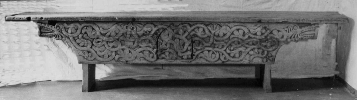 Langbord med forvaringsrom bak høye sarger. Fremsarg er med heldekkende flatskurd ranker og drageornamentikk samt en luke til forvaringsrommet. 