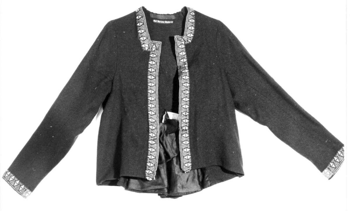 Mørkeblå trøye i ull, maskinsydd. Dekor av 3 cm brede bånd langs fremkanter, hals og ermeringning.I rygg nederst er det 3 innfeldte trekanta kiler som er foret med linlerret og bomullsstoff.