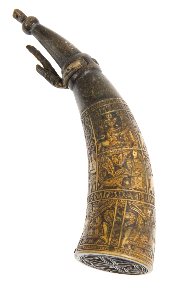 Krutthorn med flat, ornamentert bunnplate i tre. Ornamentikk på horn i form av 11 stk motivfelter omgitt av bårder med tekst. Motivene er: Adam og Eva ved Kunnskapens tre, Samson og løven, Daniel og dragen. Avbildede som rytterfigurer: Angolan, Karvel, Vidrik, Tidrik, Roland, Otvel, Burman og Olger Danske.
