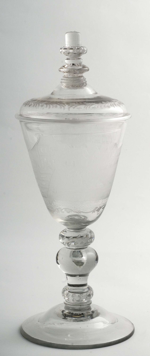 Pokal med lokk. Glass. Lokket har gravert blomsterkrans.
Balusterformet stett med dråper, cupa, traktformet, gravering i rokokko-kartusch, bilde av Bogstad før ombyggingen i 
1760-61.