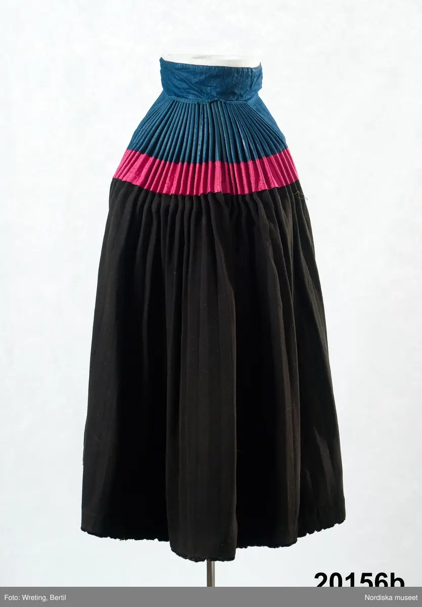 Huvudliggaren:
"Hollands draegt fr. Köpenhamn
Bestående af:
a) Kafetröje med Spaender [tillägg i blyerts.] skall vara kravetröje - 30 kr [spännet ommärkt till e+ 2011-03-08]
Spännet märkt: M.H.P.D.
b) Skjört - 28 kr
c) Forklaede med spaender - 30 kr
d) Haette med tillbehör och Haandstycke - 6 kr
e+) [spännet ommärkt till e+ 2011-03-08] Spännet märkt: M.H.P.D.
Ink. ss 20.087 [=ink. hos fru m. Wildschiödz i Köph. ss 20.001 [= gm (genom) dr A. Hazelius i Stockholm.]]- 94 kr. Under hösten 1877."

B. Kjol, "skjört"  av svart klädesberedd vadmal helfodrad med svartfärgad glättad linnelärft. Kjolkanten skodd med 4,5 cm bred svart ylleschagg. Upptill på kjolen ett 19 cm stycke av mellanblå glättad bomullslärft s.k. domestik, som bak är hårt styvat och ligger i jämna rynkor som en stel rundad platta som i nedre kanten har ett påsytt 7 cm brett rött ripsband av siden. 11 cm bred midjelinning av det blå tyget, mitt fram sprund med 3 par snöröglor av mässing i linningen.
Kjolen handsydd av material som förefaller att vara från 1700-talets andra hälft. 
Just denna typ av kjol kalls "Jöb" och bars vid sorg, Kjolen kunde då slås upp över huvudet så att den styva bakdelen fälldes upp som en solfjäder på ryggen. Se vidare Elna Mygdal: Amagerdragter, Kphmn 1930 sid 88 ff.
/Berit Eldvik 2011-11-24