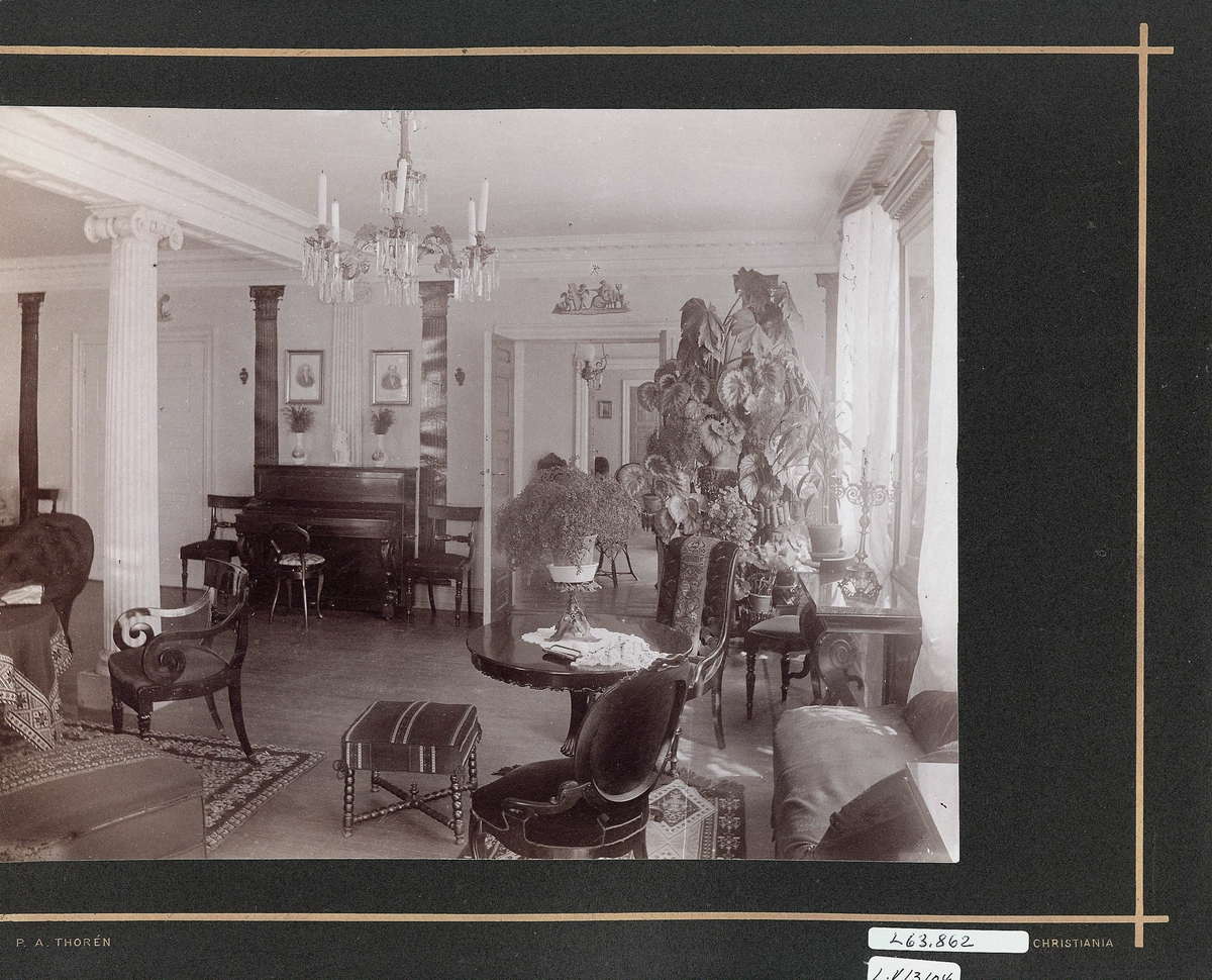 Strømsbo gård, Strømsbu, Arendal. Interiør, salen. Søyler, div. møbler som gyngestol, potteplanter, piano. Krystall lysekrone i taket. H.del av panorama, se NF.04551-015. Hovedbygningen er oppført 1760/61, men fikk sitt nåværende preg i 1804 da den ble overtatt av statshauptmann Peter Herlofson. Fredet.
Fra serie (NF.04551-001 til -021) fra billedmappen "Strømsbo", gård på Strømsbu i Arendal tilh. familien Herlofson. Bildene er fra ulike eiendommer og steder i Arendal tilknyttet familien, fotografert av P.A. Thorèn, Chra.
