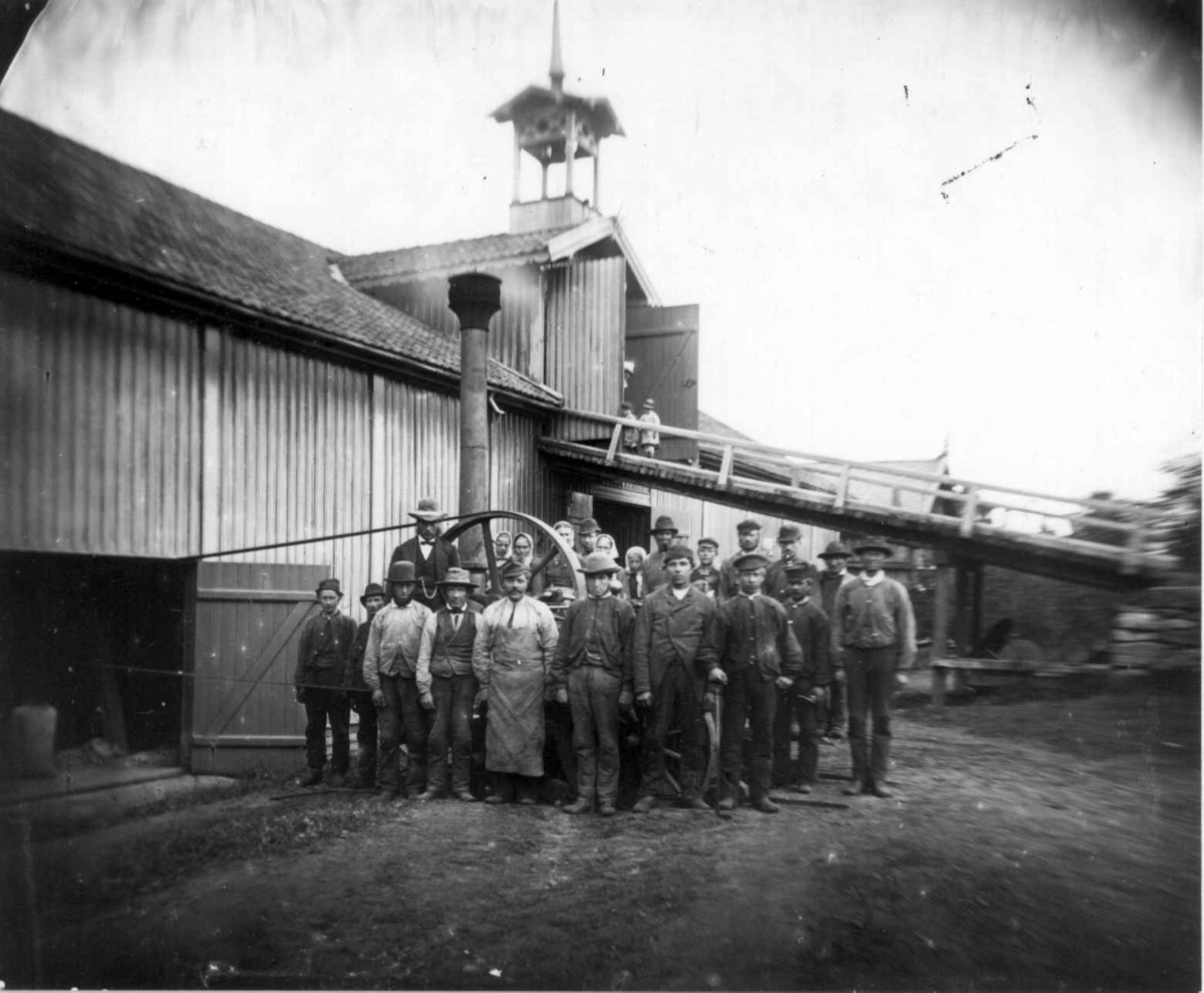 Lokomobil, Dal gård, Ullensaker. Oppstilt persongruppe ant. arbeidsmannskap til å betjene lokomobilen og treskeverket innenfor i låvebygning med låvebro og tårn. Se Andre opplysninger.
Fra serie fotografert av kammerherre Fredrik Emil Faye (1844-1903), gårdens eier.