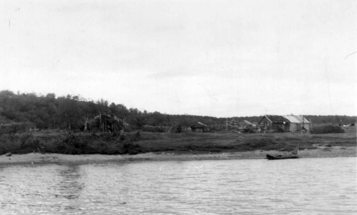 Mikkel Johansen Utsis gård med bygninger, sett fra elven, en båt ved elvebredden. Hemmujavve 1953.
