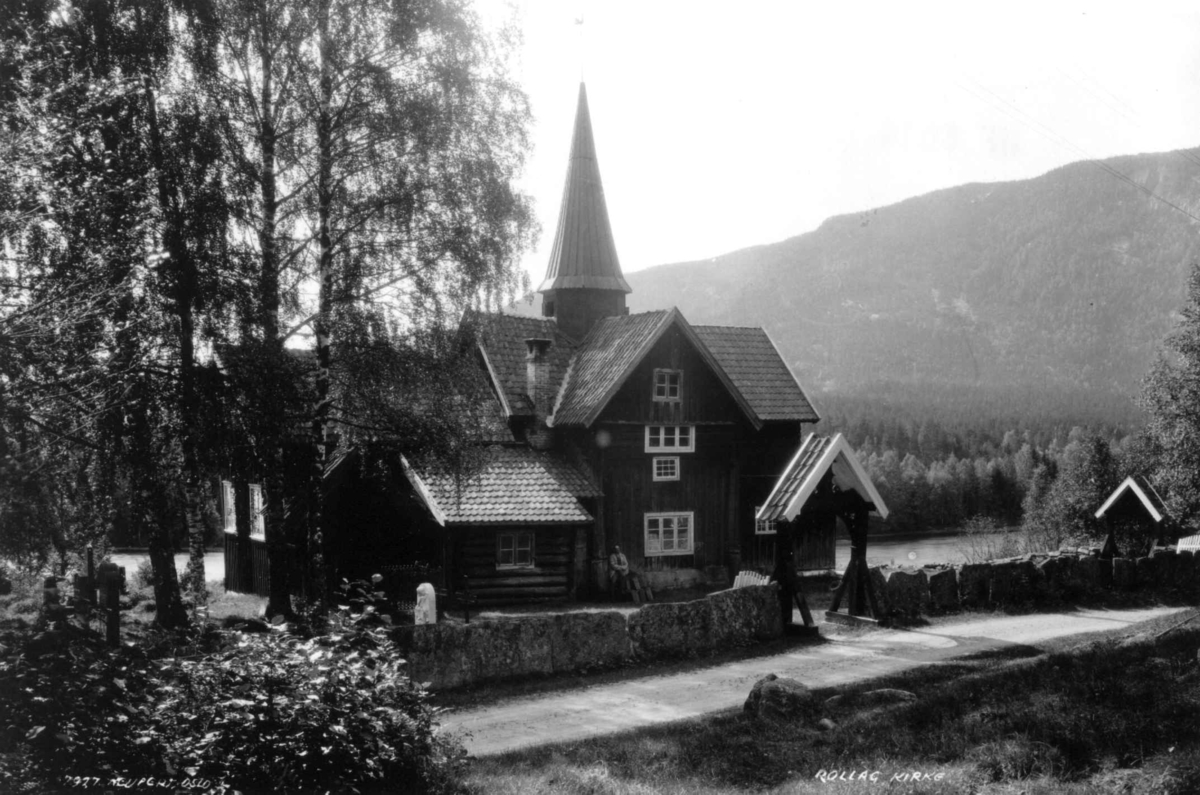 Rollag kirke - Rollag, Buskerud. 1930. Kirkegård. Trær. Gravsteiner. Mann sittende ved inngangen. Skog og åser i bakgrunnen.