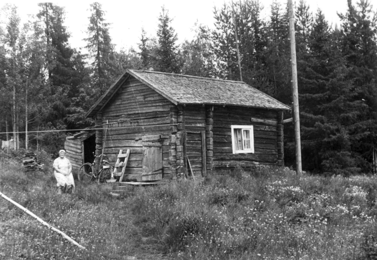 Stue/bolig bygget ca. 1850, opprinnelig inngang på langfasaden. En kvinne sitter ved huset. Olkamanki 1958.