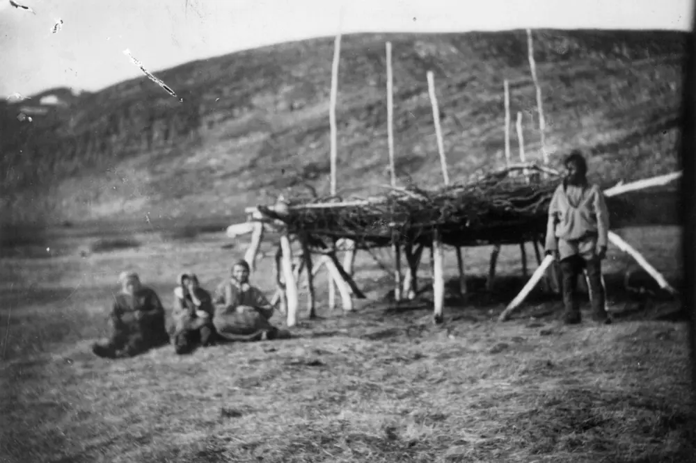 Antakelig en "luovvi", som er en samisk betegnelse på en oppbygd plass for oppbevaring. Fire personer sitter utenfor. Fra området Trollfjord og Gulgofjord.
.