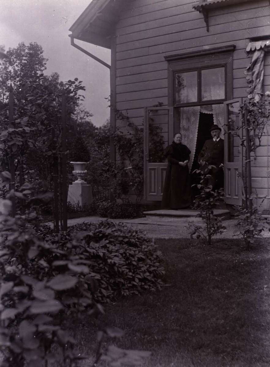 Postkort. Jule- og nyttårshilsen. Fotografisk motiv.  Svart/hvitt. Fra hage. Kvinne og mann i verandadøren. Datert 1896.