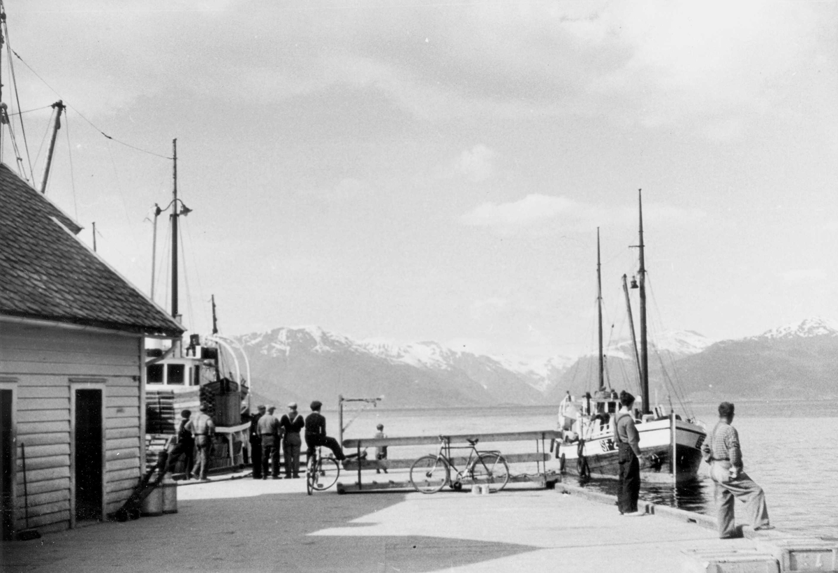 Fiskeskøyte på vei inn til brygge hvor det står arbeidsfolk.