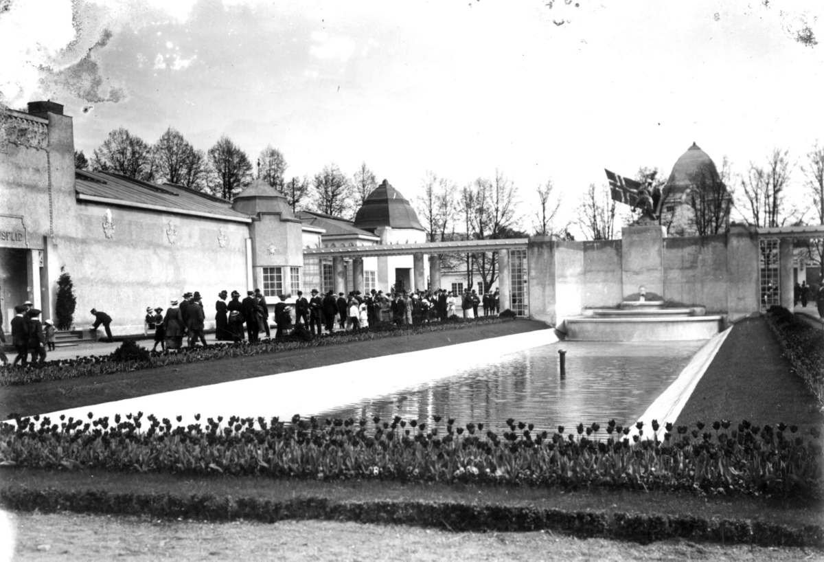 Jubileumsutstillingen på Frogner, Oslo, 1914.
Husflidsbygningen til venstre. Basseng. Skulpturen Helhesten av Gunnar Utsond i bakgrunnen. Tulipanbed i forgrunnen. Spaserende menn og 
kvinner.
kvinner.
Spaserende menn og kvinner.
