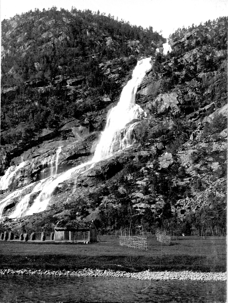 Landskap, Hildalsfoss, Odda, Hordaland. I forgrunnen kornstaur og tomme hesjer omkring et uthus. 
Del av gave fra Den norske Turistforening 1972.