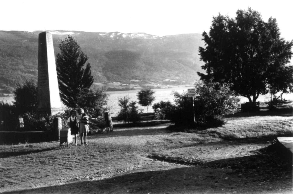 Voss 1939. Veteranbautaen omgitt av en liten park. To småjenter i forgrunnen. Vann, skog og fjell i bakgrunnen.