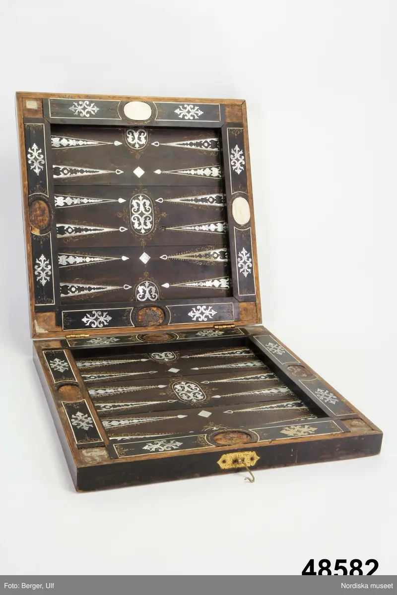 Brädspelslåda för bräde, kvarn och schack, med sju spelbrickor. Svart fanér med inläggningar av pärlemor, elfenben samt graverade förgyllda metallskivor. Blindträ av ek. (Uppgifter från katalogkort). Tillhörande träask för förvaring av spelpjäserna.