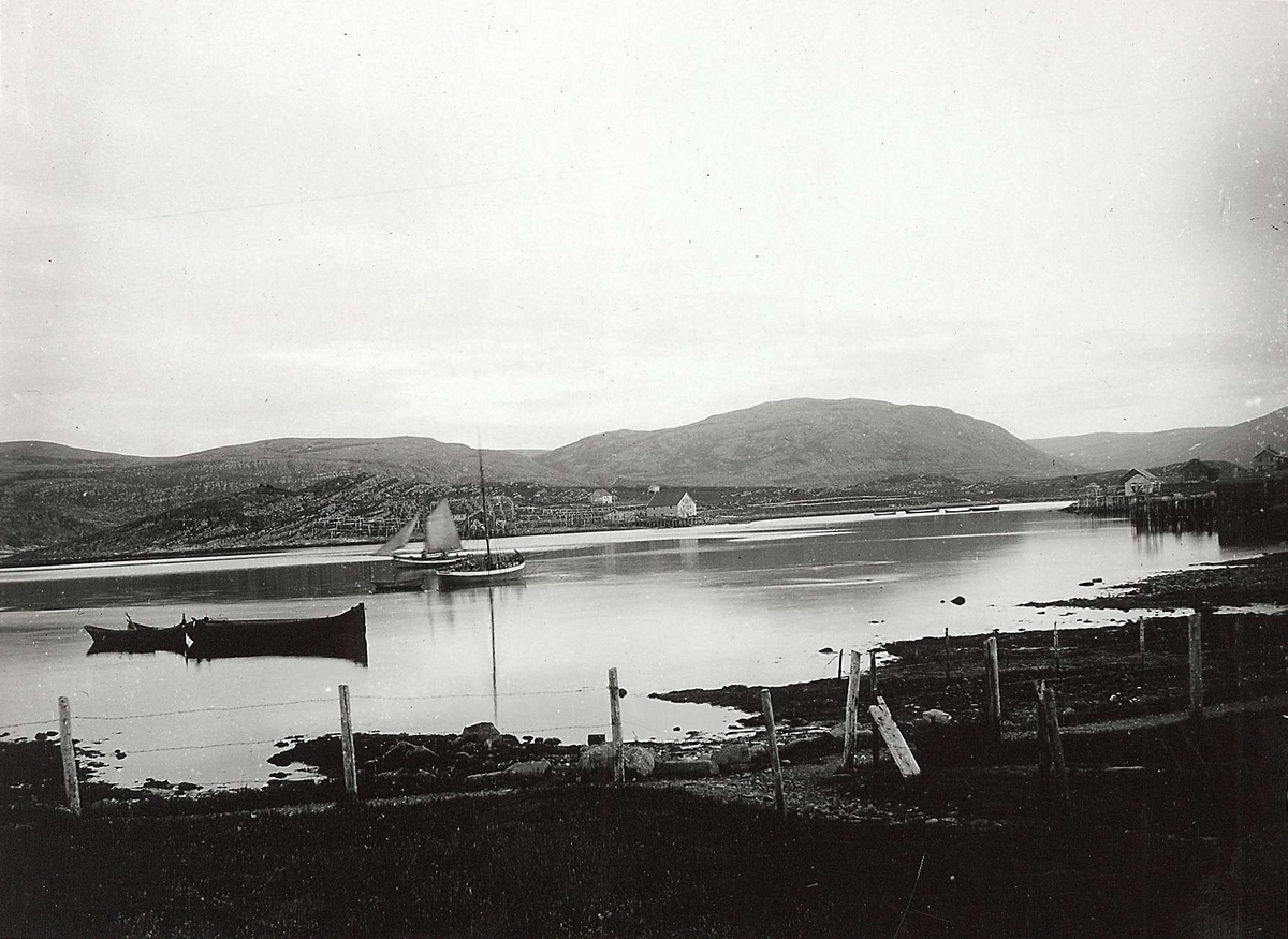 Landskap, Båtsfjord, med utsyn over fjorden med ulike båter, noen under seil. Noe bebyggelse, med fiskehjell i bakgrunnen. Del av serie fra en forskningsreise i Øst-Finnmark 1909. 