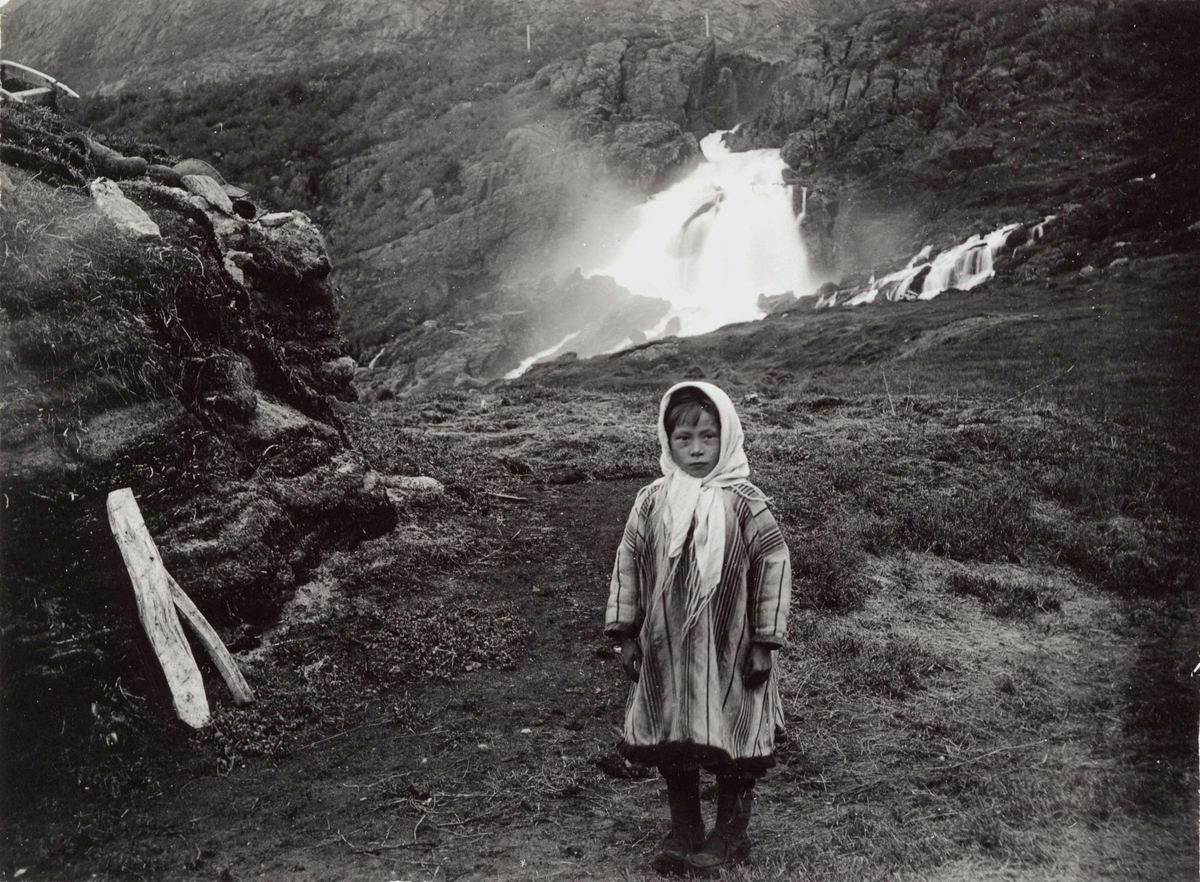 Familien Johansen i samisk drakt foran gamme ved Adamsfjordfossen i Lebesby. Piken til høyre er Valborg.
Del av serie fra en forskningsreise i Øst-Finnmark 1909.