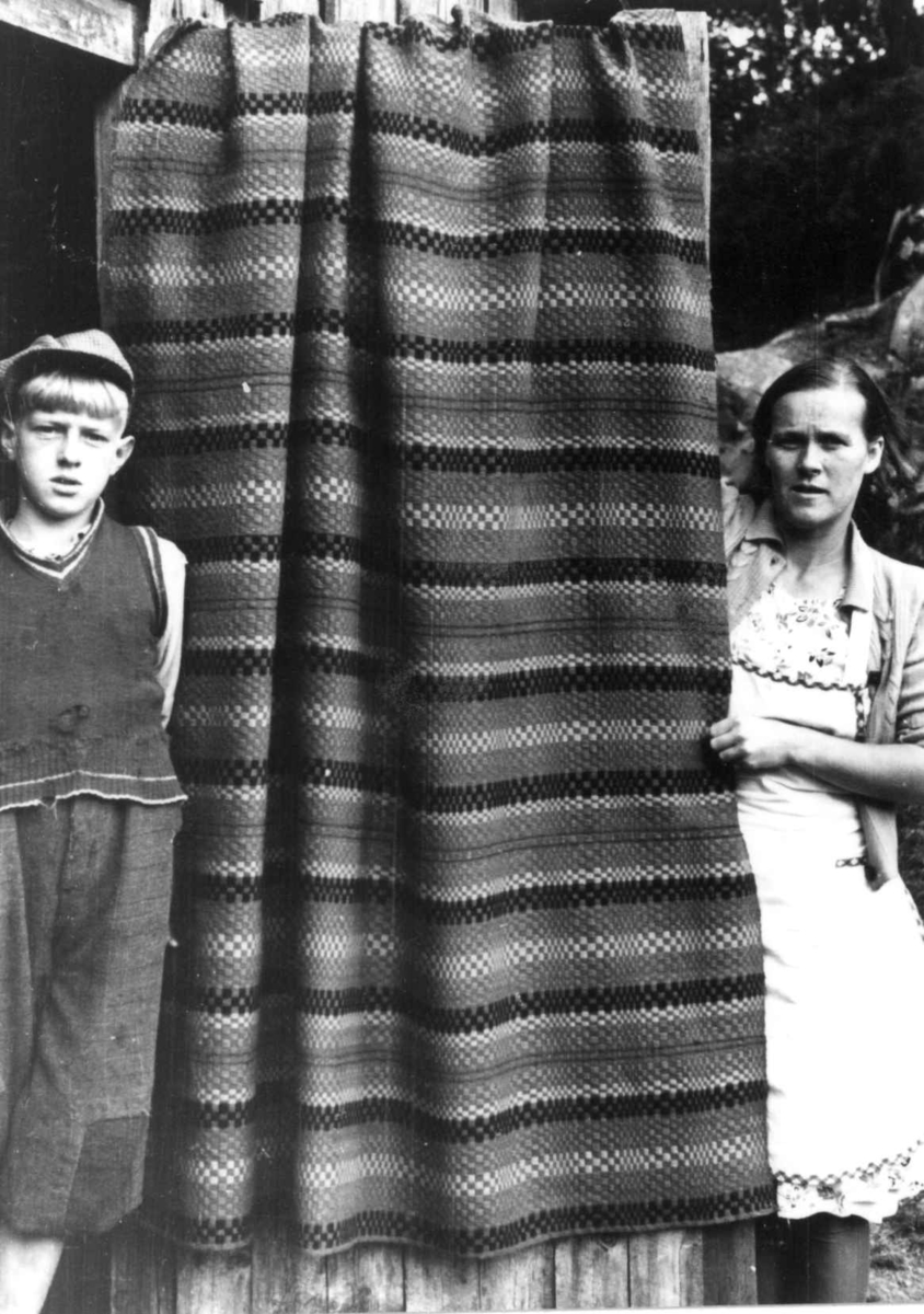 Iveland, Vest-Agder 1941. Teppe, muligens tavlebragd, spunnet og vevd ca. 1900. To personer står ved siden av det hengende teppet.
