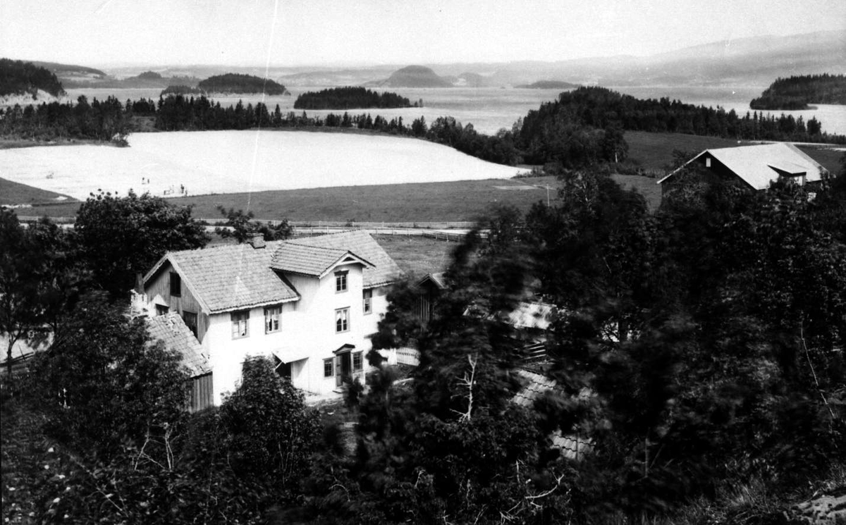 Utsyn over landskap ved Sundvollen, Hole/Ringerike, Buskerud, ant. med Tyrifjorden i bakgrunnen. I forgrunnen bolighus på gård. Deler av Brødrene Brunskows serie norske landskapsbilder.