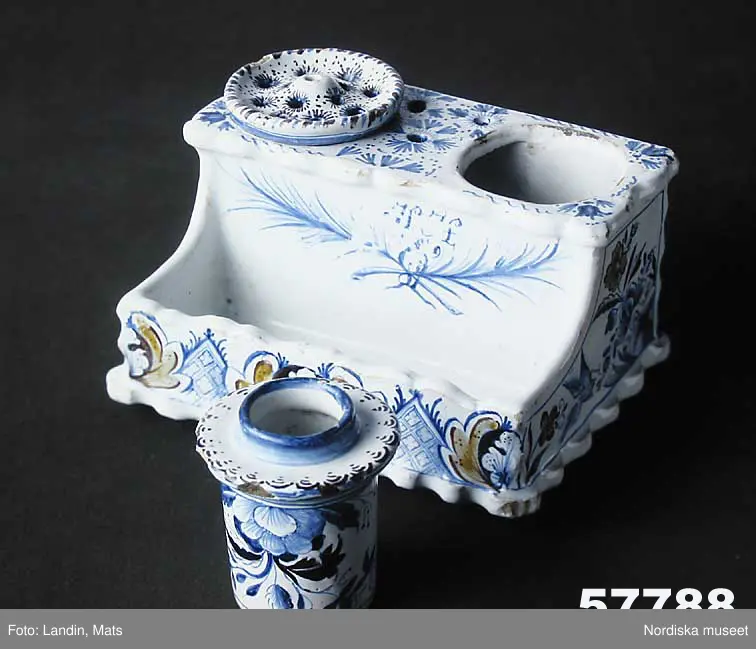 Katalogkort:
"Skrivtyg, fajans. Rektangulärt, framtill rum för pennor, upptill bläckhorn och sanddosa; dekor. med blommor och rutmönster i blått, brunt och gult, på framsidan två palmkvistar i blått, varöver H.H. 
Sign. i blått: Stockholm 2/8 1749 Hindrich Höök.
/Upmark 1904"

Skrivställ av fajans med dekor i blått och manganviolett, Rörstrand 1749.
Skrivstället med runda dosor för bläck och strösand är målat av Henrik Höök som signerat det med hela sitt namn under botten, oftast står bara målarens initialer utsatta. Han var då lärgosse vid fabriken och blev målargesäll 1754. 
Märkning: "Stockholm 2/8 1749" samt "Hindrich Höök".
Byggmäst E A Bomans samling nr  75.
/Annika Tegnér 2006