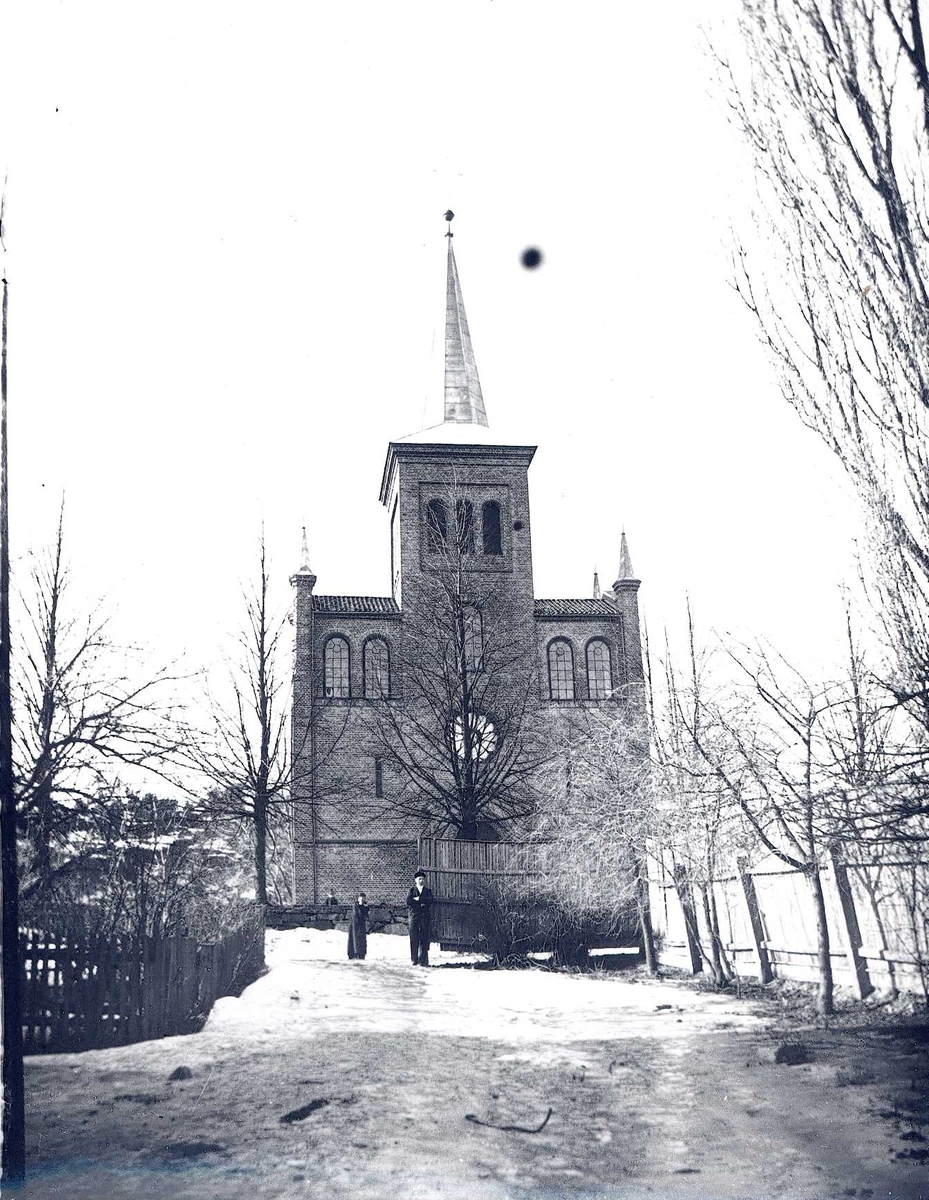 Vei med gjerder går mot kirke. Spist tak på tårnet, små tårn på "skuldrene".