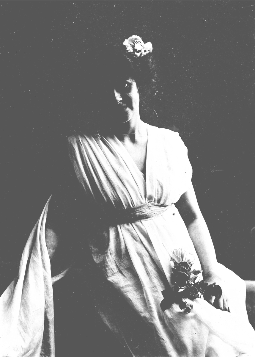 Studioportrett av kvinne i kostyme med hvit kjortel og blomster i håret. I den ene hånden holder hun en blomsterbukett.