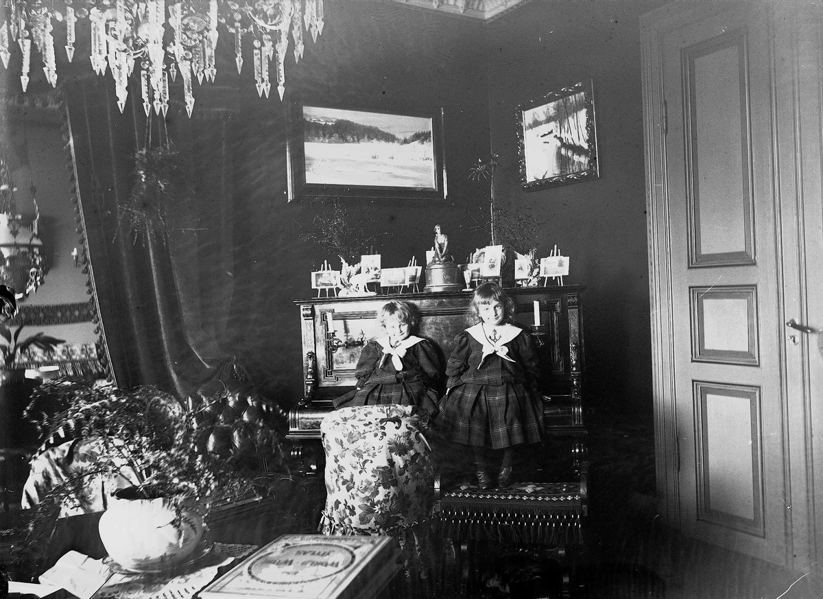 Gudrun og Karen Q. Wiborg sitter på piano i stue, Meltzers gate 9, Oslo, 1899. Fotografier i ramme plassert på pianoet.