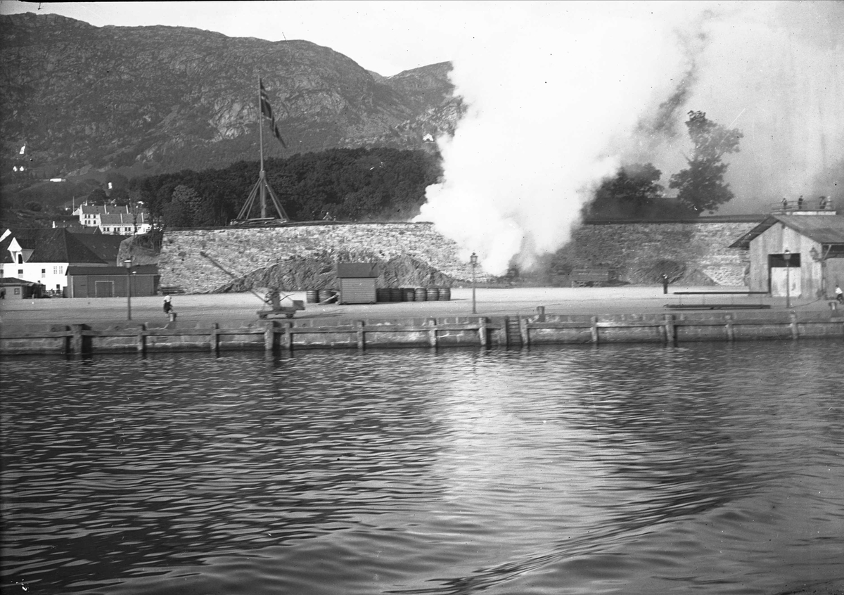 Båter, havneanlegg og bygninger. Flagg. By ved kysten mot fjellet, tatt fra båt. Bergen? Bilde fra reise med båt til Trondheim. 1902.