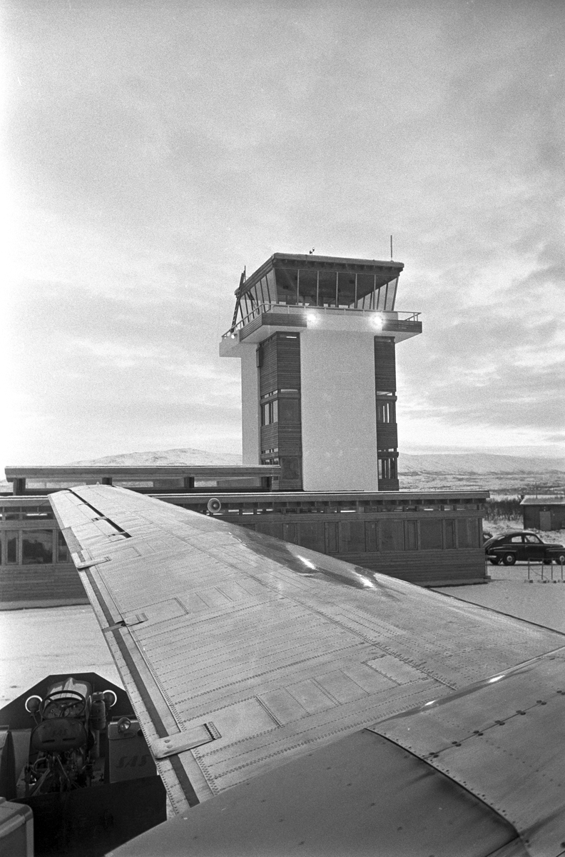 Serie. Ombord på SAS flyet til Tromsø. Flyfoto over snødekkete fjell. To flyvere i flytårnet. To menn og en kvinne, ant. Eva Lie, sitter rundt et bord. Fotografert desember 1965. 