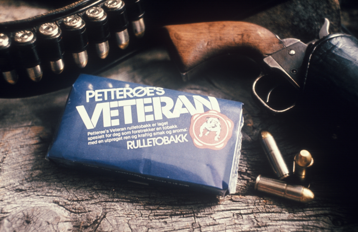 Reklamefoto. En pakke tobakk av typen Petterøes Veteran sammen med revolver og ammunisjon. Kampanje for tobakksmerket Petterøes Veteran 1975.