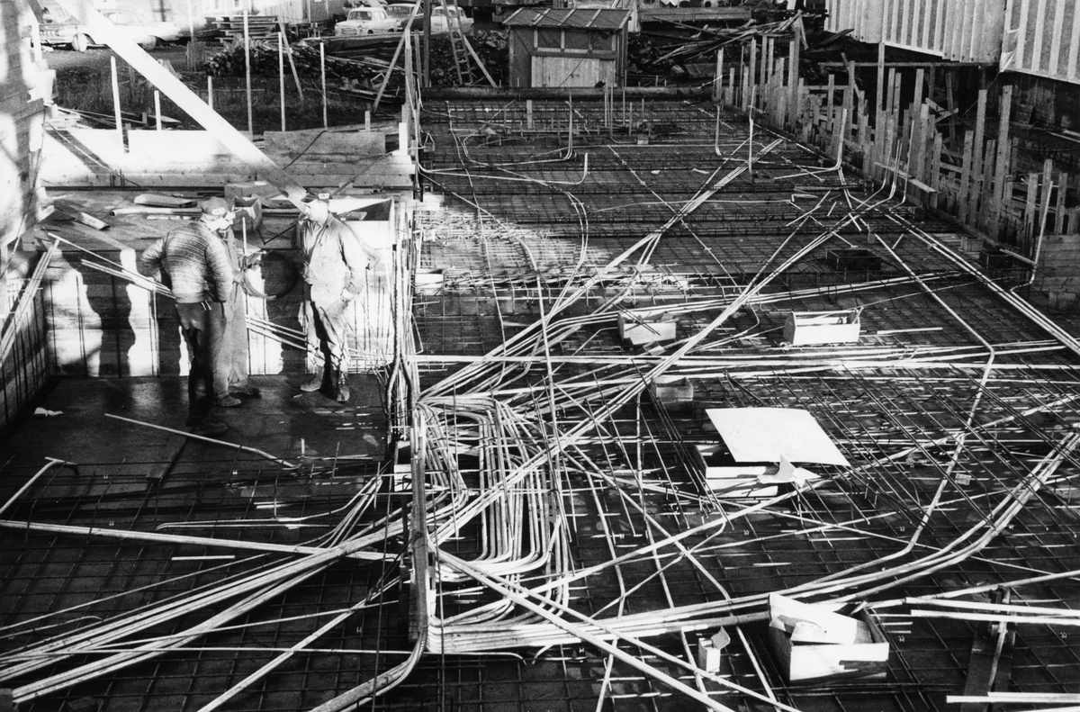 Byggeplass.
Konstruksjon av Tiedemanns Tobaksfabrik på Hovin i 1967. Tre stk bygningsarbeidere står på byggeplassen å snakker.