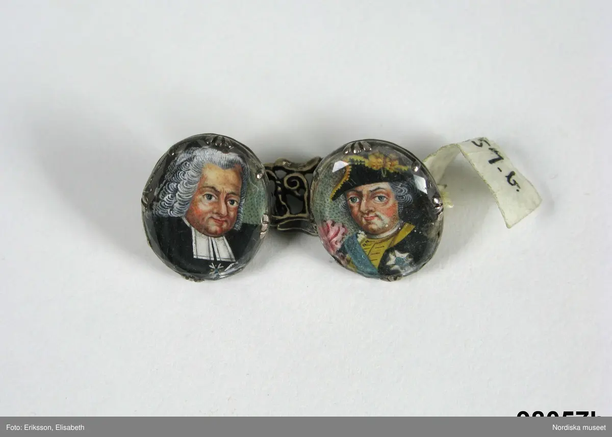Manschettknappar, två par, miniatyrer. 1700-tals mitt.  Silver, glas.
Saknar stämplar. På varje knapp ett ovalt porträtt i miniatyr i silverinfattning, baksidan räfflad, knapparna förenade två och två, på den ena föreningslänken. ”I H” i genombrutit arbete, den andra föreningslänken senare ersättning. Manschettknapparna hölls i ett rött marokäng (getskinn) etui.
(katalogiseringen gäller a-c)