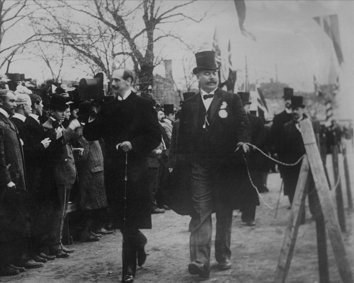 Avfotografering.
Gruppeportrett, kong Haakon VII hilser tilskuere med hatten.