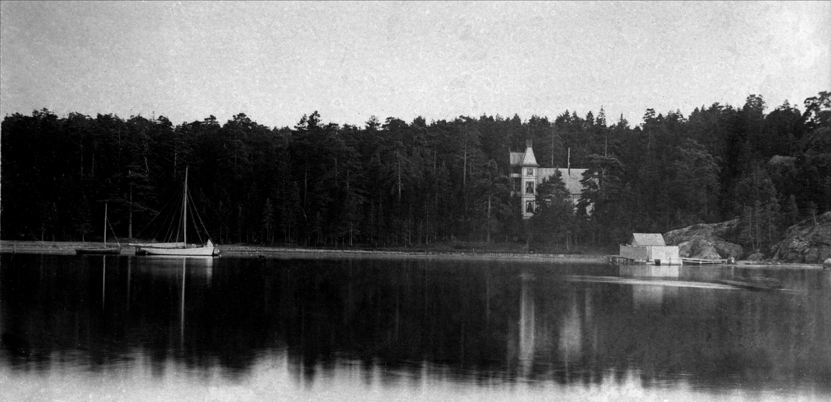 Landsted ved vannet, seilbåt, brygge og stor bolig i vannkanten.