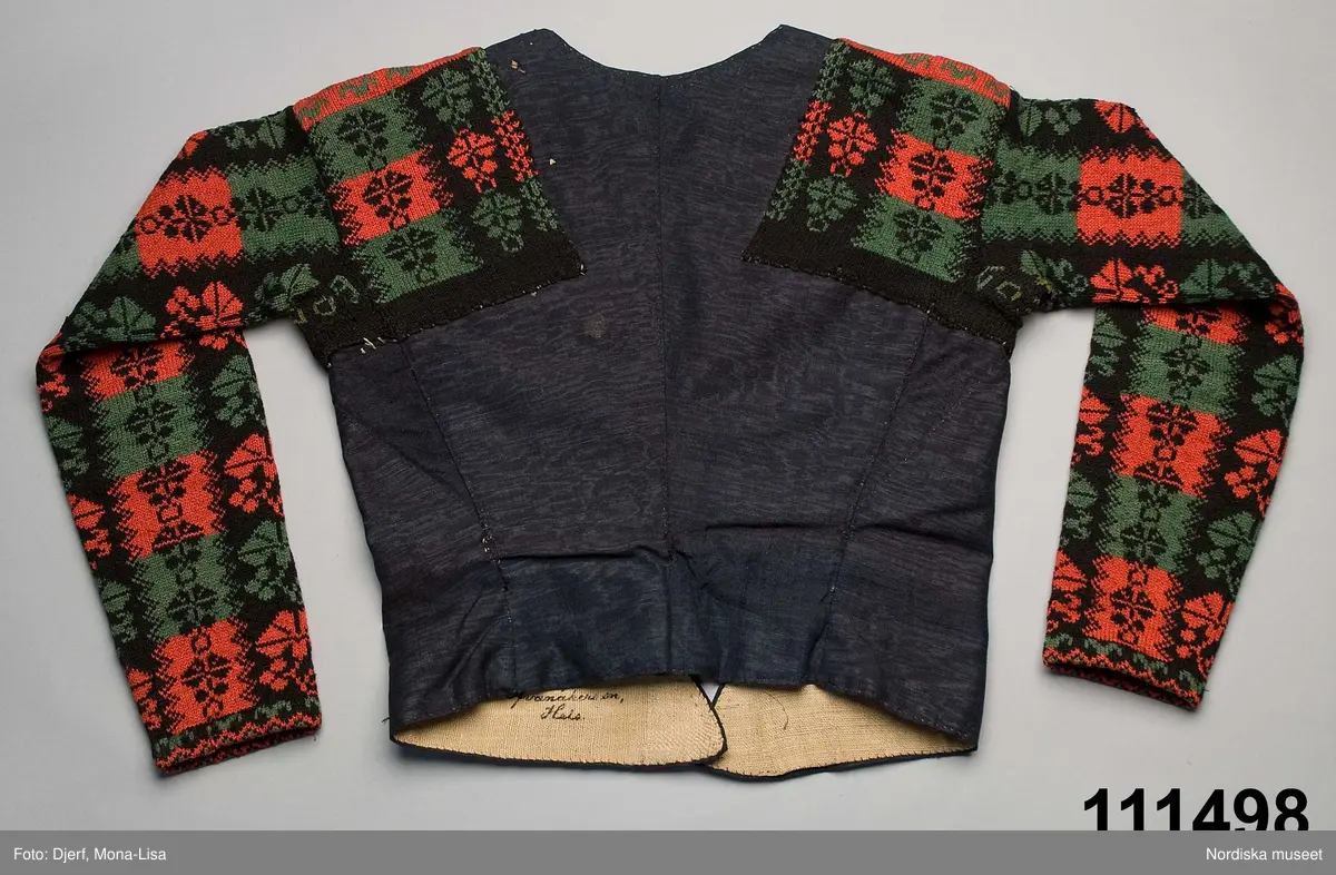 Ursprungligen ett livstycke av mörkblått kamgarnsylle i tuskaft med vattrad yta, s.k. kamlott. Livstycket i 1700-talssnitt med långt skört.
2 fram-och sidstycken, 2 bakstycken med 3 skörtveck i ryggen, 
Knäppning med 6 par hakar och hyskor, de av järntråd är äldst och ursprungliga, de i mässing yngre. Foder av grov oblekt linnelärft. Vid omsyning till tröja har skörtvecken sytts in i sömmarna ev. har hela livstycket även kortats något. Ärmarna mönsterstickade av rött tvinnat ullgarn och vitt bomullsgarn i stora rutor med nejlikmönster instickade i motsatsfärg. Ett rakt axelstycke stickat i samma mönstring och fastsytt vid ärmen och därefter påsytt på livstycket, så att det ursprungliga ärmhålet finns kvar under det stickade.
Anm. Ärmarna har fållats upp ca 2 cm

Kamlott var modetyg under senare delen av 1700-talet, det var ett dyrbart köpetyg och användes av allmogen i de finaste bröllopsplaggen som livstycken och kjol. Se t.ex svart kjol och tröja från Arbrå inv.nr 1824 och 1825. Ofta ser man sådana plagg omsydda till modernare snitt under 1800-talet. Ett så dyrbart och fint tyg återbrukades gärna. Jfr. liknande tröjor inv.nr 111497 och 201427 samt lösa ärmar 113011.
Troligen är samtliga dessa lösärmar stickade av samma person.
Beskrivning finns i boken Svenska Tröjor av Britt-Marie Christoffersson, ICA förlag 1988.
/Berit Eldvik 2006-06-16