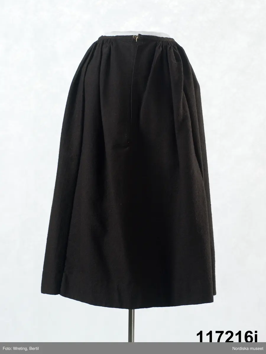 Huvudliggaren:
"Sorgdräkt för kvinna. Särk med 'hängseltåg', 'särkyvdel'; vit -'häsklä'; blått livstycke; svart tröja; huvudduk; 'svart näsduk'; hatt; 'raskförklä', 'sockar',  'hårbandsvifsa', huvudbindel."

Bilaga:
a: "8 Särk, övdel. av linne eller blågarn. med broderade linningar på överdelen"
c: "11. Hvit häsklä."
Anm: Kung Kerstin Larsdotters resa i Floda sn Dalarna 24/2 - 8/3 1912. Innehåller förteckning på inköp av dräktdelar från Floda, inv nr 117 216 - 117 239. Här finns  tillverkningstid och beskrivningar på vissa av dräktdelarna.  I vissa fall nämns också utförligt på vilka helgdagar dräktdelen  användes.

Katalogkort:
a: Hängselsärk
b. Överdel av halvlinne vävt i kypert.
c.  Halskäde av småmönstrat bomullstyg, liknande kambris, vävt  i kypertvariation med drällmönster.
d. Livstycke av mörkblå kraftig vadmal,
e. Tröja av grov svart vadmal

i. 
 Mönstervävt band med upplockat mönster i rött z-tvinnat ullgarn på vit linnebotten, med hjärtan, kryss och rombformer, 2 band om vardera 124 cm har vikts och sytts ihop så att det ena bildar en ögla och det andra har fria ändar avslutade med en tofs i bandets garn.
Läs mer om dräktskicket i Floda i: Albert Alm, Dräktalmanacka för Floda socken, Nordiska museet, Sthlm 1930
/Berit Eldvik 2005-04-14