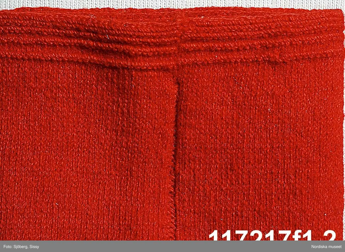 Huvudliggaren:
"Brudpigedräkt, särk med "hängseltåg", "yvdel", röd kjortel, kattunsförklä, silkeshalsklä, röda "sockar", "hårbandsvifsa", huvudbindel."

Utdrag ur bilaga förteckning:
"25. Kartunsförkläde. sen 1700 talet
26. Röd kjol. av kläde. bars av brudar som inte hade krona, brudpigan blå, vid kronbrudar bars den af brudpiga och även på juldagen, Påskdagen, Pingstdagen.
27. Blå kjol. av handspunnet garn, hemväft, gjort 1875
28. Silkeshalskklä. som även bars av brudar, och brudpigor"
"31. Bindel som bars av brudar  Brudpigor, av silke  1700"

Katalogkort:
A. Finns ej.

A. Hängselsärk
B. Överdel, "yvdel" 
C.
D.
E.
F.
G.
H.

A: Längd: 86 cm. Längd: 20 cm - livdel. Längd: 42 cm - axelband. Bredd: 1 cm - axelband.  Vidd: 132 cm. Vidd: 88 cm - midjevidd.
Hängselsärk med ihopsydd liv- och nederdel. Rynkningar i sidorna fram och mitt  bak mot livdelen. Rak livdel av kypertvävt bomullstyg, av två stycke, varav ett är ett 30 cm brett mittstycke. Två fastsydda axelband, ripsvävda i bomullsgarn. Nederdel av kypertvävt halvlinne, bomull i varp och lingarn i inslag, sidsömmar med 7 cm långa sprund. Alla sömmar utom sammanfogningssömmen mellan liv- och nederdel är maskinsydda.
/Inga-Lill Eliasson 2006-11-14 

B: längd 37 cm, längd mellan ärmlinningarna 165 cm, ärmvidd 50 cm.
Överdel av bomullslärft, handsydd, ärm och liv i ett stycke med skarvremsa fram i nederkant. Halssprund och -ringning med smal fåll. Ärmlinning med rätlinjigt broderi i vitt lingarn.
Berit Eldvik 2004
